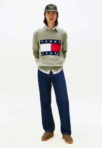 Pull léger vert avec le logo "TOMMY JEANS", manches côtelées, superposé à une chemise à col, associé à un pantalon bleu foncé et des chaussures brunes.