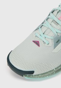 Light gray athletic shoe with a textured mesh upper, teal laces, pink accents, and a clear sole featuring green speckles.