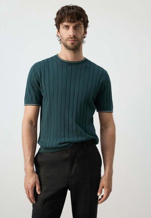 Homme aux cheveux courts et bouclés portant un t-shirt côtelé vert foncé à manches courtes et un pantalon noir, debout devant un fond uni.