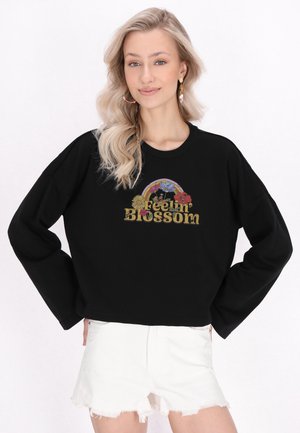 Młoda kobieta z długimi blond włosami w czarnej bluzie z kwiatowym napisem „Feelin' Blossom” i białych, postrzępionych szortach jeansowych.