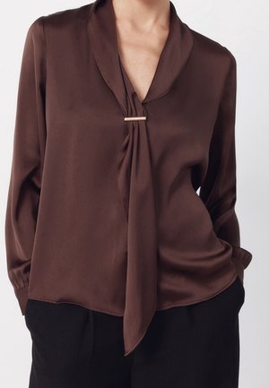 Femme portant un chemisier marron à manches longues avec un détail noué sur le devant et un pantalon noir, mains dans les poches.