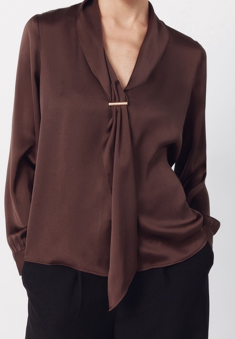 Femme portant un chemisier marron à manches longues avec un détail noué sur le devant et un pantalon noir, mains dans les poches.