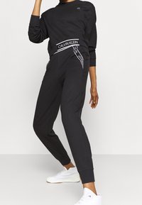 Sweat-shirt noir et joggings en tissu doux. Les joggings disposent d'une ceinture à logo et de poches latérales. Assorti à des baskets blanches.