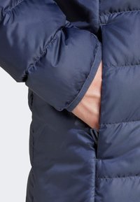 Marineblå oppustelig jakke med quiltet design, der har en skjult lomme og teksturerede ærmekanter. Lavet af letvægts, vandafvisende materiale.