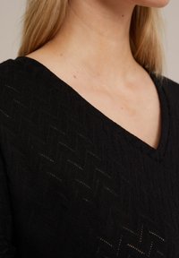 Čierny pletený top s výstrihom do V, textúrovaným herringbonovým vzorom a polotransparentným povrchom, ktorý zvýrazňuje zložitú štruktúru látky.