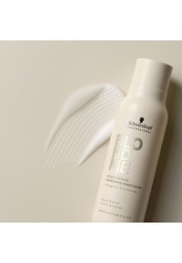 Conditionerfles met een beige, matte afwerking, zilveren opdruk; crèmige textuur verspreid ernaast, met een golvend patroon. 250 ml formaat.