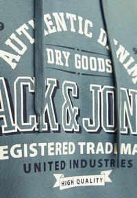 Grøn hættejakke med hvid og navy grafisk tekst, der inkluderer "AUTHENTIC DENIM" og "JACK & JONES", med en flettet snor og blød tekstur.