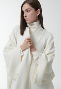 Witte geribbelde stofomslag met lange mouwen, featuring een hoge kraag en gedrapeerd ontwerp. Model houdt de stof dicht bij zich, waarmee de textuur wordt getoond.