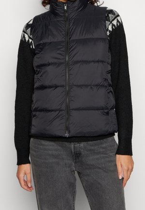 Person iført en sort quiltet vest med lynlås over en sort sweater med hvide mønstre på skuldrene og mørkegrå jeans.