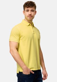 Gul polo skjorte i bomuldsblanding, korte ærmer, foldet krave og sidelæg i kanten. Modellen har mørke jeans på.
