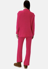 Roze ribgebreid gebreid trui- en broekset, met lange mouwen, een hoge halslijn en wijde broekspijpen. Het materiaal lijkt zacht en textuur te hebben.