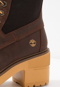 Hnědé kožené kotníkové boty s texturovaným černým elastickým panelem, zlatým logem a odolným žlutým gumovým podpatkem s texturovaným povrchem.