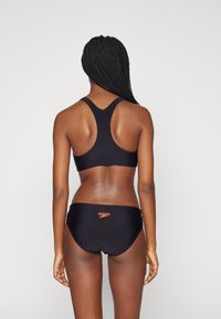 Baño negro con parte superior de espalda nadadora y braguitas. Presenta una tela suave, costuras mínimas y un logo naranja en contraste en la parte posterior.