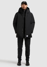 Svart vadderad jacka med huva, med en dragkedja fram, bärs över en svart polotröja, i kombination med mörka jeans och svarta stövlar.