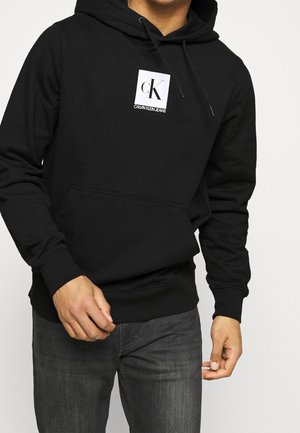 Mand iført sort Calvin Klein-hoodie med hvidt logopatch og mørkegrå jeans, stående foran en ensfarvet baggrund.