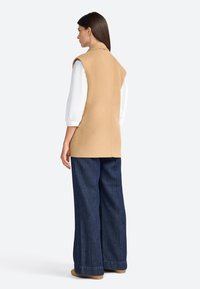 Beige mouwloze vest met zij-splitten, gecombineerd met oversized donkerblauwe denim broek, witte wijdvallende mouwen en lichtbruine schoenen.