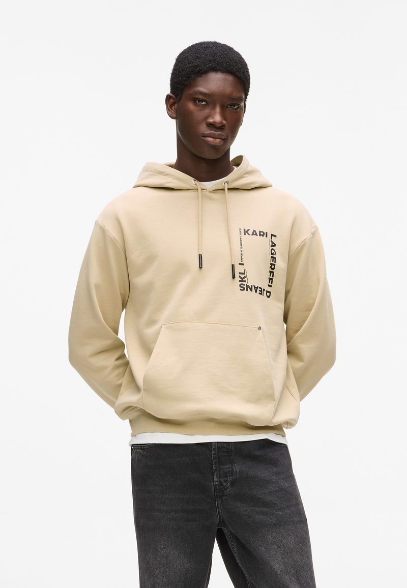 Felpa con cappuccio beige in misto cotone, con tasca a marsupio frontale e testo stampato in nero sul lato sinistro. Jeans scuri completano il look.