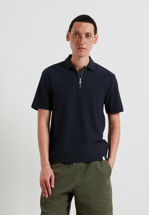 Polo a maniche corte blu navy con cerniera frontale, texture a costine e colletto piatto, abbinato a pantaloni verde oliva.