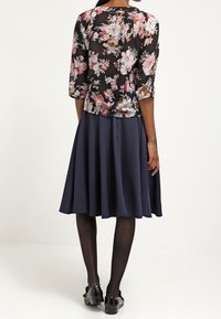 Blusa nera a fiori con maniche tre quarti, abbinata a una gonna svasata blu navy. Il modello indossa collant neri trasparenti e scarpe nere.
