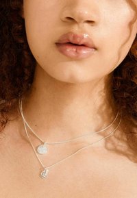 Glanzende lippen en krullend haar omlijsten een nek met daarop twee delicate zilveren kettingen met ronde hangers.