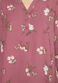 Paarse blouse met een V-hals en knoopdetails, versierd met bloemen- en vlinderpatronen in roze en groen. Zachte stof met een vloeiende pasvorm.