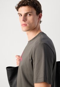 Majica kratkih rukava u maslinastoj boji s roza grafičkim otiskom, sadrži okrugli izrez, mekani materijal i vidljive šavove.