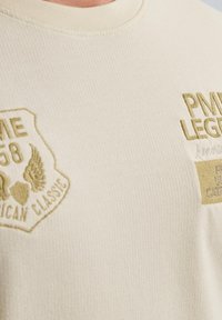 Jasno kremowy t-shirt o fakturowanym materiale z prążkowanej tkaniny. Posiada haftowane łatki w złotym kolorze, w tym tekst i grafikę z przodu.