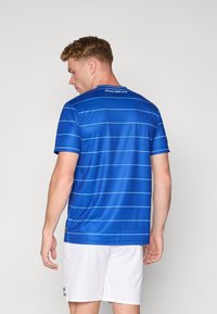 Maglia atletica blu con strisce orizzontali azzurre, maniche corte, scollatura rotonda e texture del tessuto. Indossata con pantaloni corti bianchi.