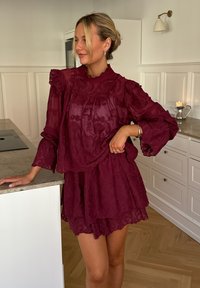 Blouse en dentelle bordeaux avec un col et des manches volantés, associée à une jupe à volants assortie ; texture transparente avec des détails de motifs floraux.