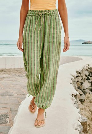 Person läuft auf einem geschwungenen Küstenweg, trägt grün gestreifte, weite Hose und beige Flip-Flops, Ozean und Himmel im Hintergrund.