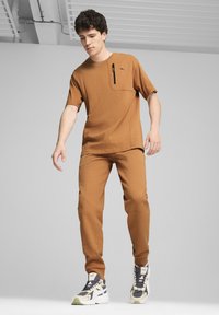 Hnedé športové tričko so zipsovým vreckom, skombinované s do seba ladícími hnedými teplákmi. Outfit sa vyznačuje voľným strihom a hladkou textúrou látky.