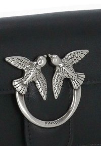 Borsa a tracolla in pelle nera con un accento in argento raffigurante due uccelli che si fronteggiano, ali dettagliate e un anello metallico circolare.