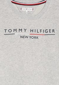 Harmaa collegepaita, joka on valmistettu puuvillasekoitteesta. Paidan kauluksessa on ribbireunus, ja siinä on rohkea "TOMMY HILFIGER NEW YORK" -logo sekä korostusrivit punaisena ja laivastonsinisenä.