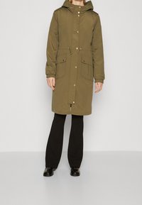 Olivgrüner Parka mit Kapuze, vorderen Knöpfen, Taillen-Zugband und zwei großen Taschen. Hergestellt aus glattem Stoff mit elastischen Bündchendetails.