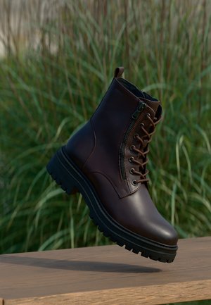 Dunkelbrauner Leder-Schnürstiefel mit seitlichem Reißverschluss und dicker schwarzer Sohle, schwebend über einer Holzoberfläche mit grünem Gras im Hintergrund.