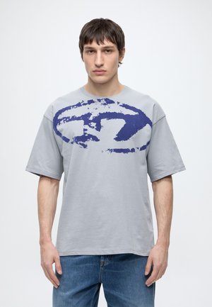Jeune homme portant un t-shirt gris clair oversize avec un grand motif abstrait bleu foncé et un jean bleu, debout devant un fond uni.