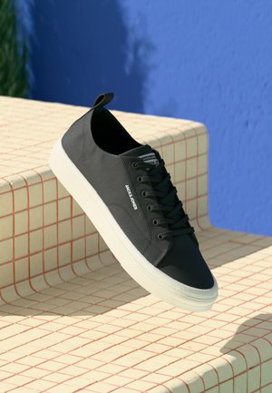 Black leather sneakers with a white rubber sole, featuring black laces, a pull tab, and a stitched side logo on a patterned surface.