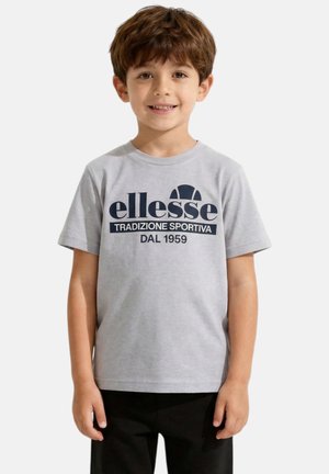 Garçon aux cheveux bruns portant un t-shirt gris clair avec le texte « ellesse TRADIZIONE SPORTIVA DAL 1959 » et un pantalon noir, debout et souriant.