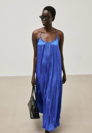 Frau trägt ein fließendes blaues Satinkleid in Maxi-Länge und schwarze Sonnenbrille, hält eine schwarze Handtasche vor einem einfarbigen Hintergrund.