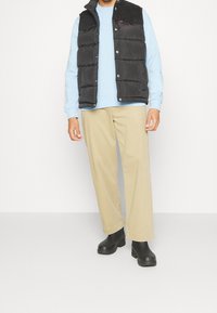 Gilet imbottito nero con colletto in velluto a coste, indossato sopra una felpa azzurra chiara, abbinato a pantaloni beige a gamba larga e zoccoli neri.