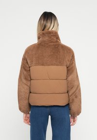 Smeđi kaput od umjetnog krzna s podstavljenim, glatkim donjim dijelom. Visoki ovratnik i oversized rukavi. Jednostavan dizajn, bez vidljivih metalnih dijelova ili uzoraka.