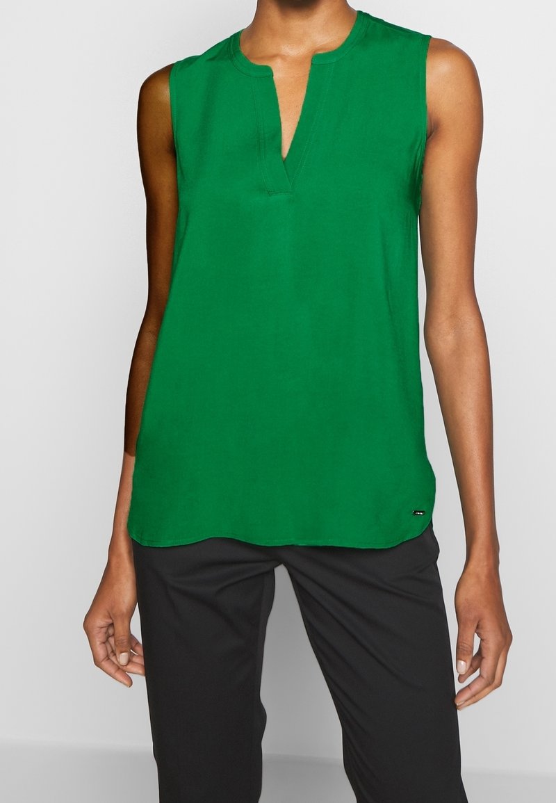 Green sleeveless top featuring a v-neckline and a curved hem. Made of smooth fabric with a subtle sheen, paired with black pants.