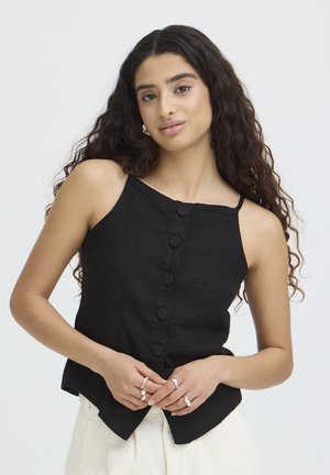 Femme aux longs cheveux bouclés portant un haut noir sans manches à boutons et un pantalon blanc, montrant des bagues en argent sur ses doigts sur un fond clair.