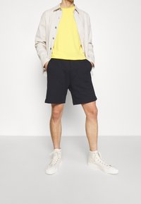 Veste boutonnée beige clair, t-shirt jaune, short bleu marine foncé et baskets montantes blanches. Design simple avec une coupe décontractée.