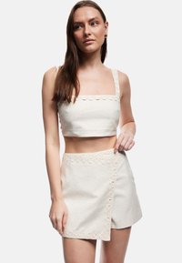 Młoda kobieta ubrana w białą bezrękawnikową bluzkę crop top i pasującą spódnicę z koronkowym wykończeniem, stojąca na jednolitym jasnoszarym tle.
