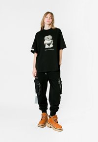 Camiseta negra de gran tamaño con un estampado gráfico, combinada con pantalones cargo negros con bolsillos con cremallera y botas marrones con cordones y suela gruesa.