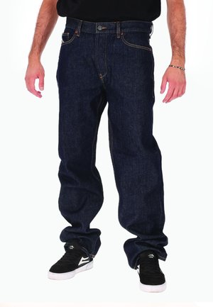 Uomo che indossa jeans larghi blu scuro e sneakers nere, in piedi davanti a uno sfondo bianco, con le braccia rilassate lungo i fianchi.