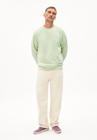 Sweatshirt vert clair, pantalon large crème et baskets violettes. Le sweatshirt a des poignets côtelés, et le pantalon présente des détails de couture visibles.
