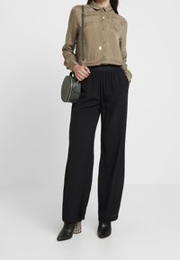 Satinová olivově zelená košile na knoflíky, černé široké kalhoty, texturovaná kulatá zelená crossbody taška a černé kotníkové boty na podpatku s akcentními detaily.