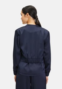 Veste bomber en satin bleu marine avec taille élastique et poignets élastiques. Texture lisse, design simple et détails minimalistes, présentée de dos.
